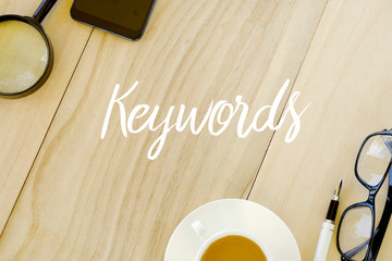 Top view of magnifying glass,mobile phone,a cup of coffee,pen and glasses on wooden background written with Keywords.