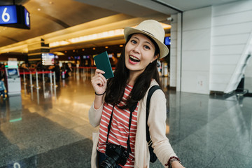 Happy young asian woman casually dressed standing in airport lobby terminal gate holding photo camera showing passport with flight tickets. travel girl in hat on summer vacation going abroad.