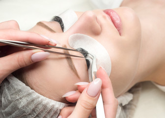Eyelash extension procedure. Woman face and experts fingers with tweezers, close up, selective focus