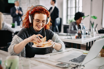 Happy redhead businesswoman eating a sandwich at her office desk. - Powered by Adobe