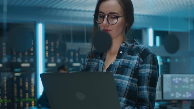 Portrait of a Smart Focused Young Woman Wearing Glasses Holds Laptop. In the Background Technical Department Office with Specialists Working and Functional Data Server Racks