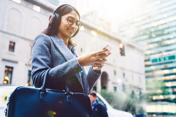 Smiling Indian female listening to music in headphones while standing on city street and relaxing after work. Young woman enjoying playlist in earphones. Watching content in social network, millennial