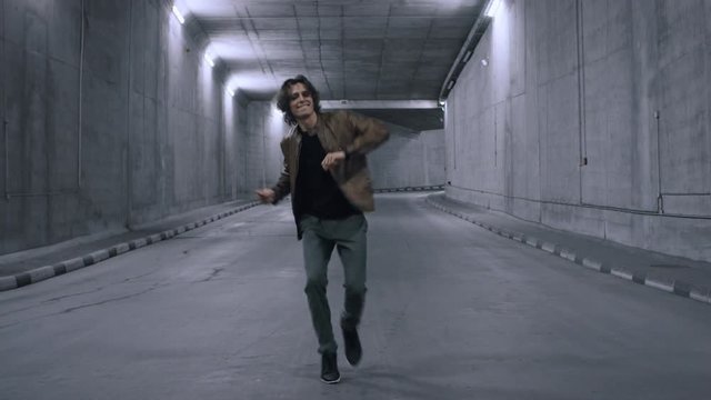 Serious Cool Young Hipster Man with Long Hair is Energetically Dancing Hip Hop in a Lit Concrete Tunnel. He's Wearing a Brown Leather Jacket.