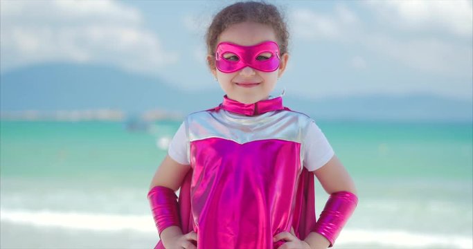 Beautiful Little Girl in the Superhero Costume, Dressed in a Pink Cloak and the Mask of the Hero. Plays on the Background Sea and Blue Sky and Clouds, Sends a Fist Forward. Concept of a Happy