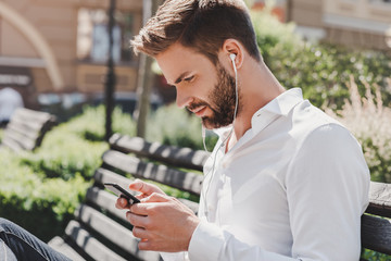 Young smiling man sitting on bench in park, listening to music with earphones on smartphone. Rest, education and relax concept