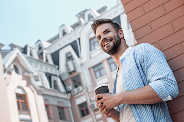 Fototapeta premium Success is peace of mind. Portrait of confident brown-haired man holding coffee cup and smiling aside while walking outdoors