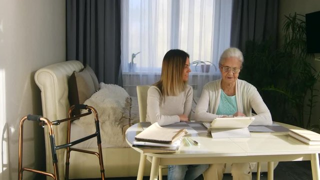 Tracking shot of senior woman in glasses sitting at table in the bedroom and reading something on screen of digital tablet with help of young female volunteer