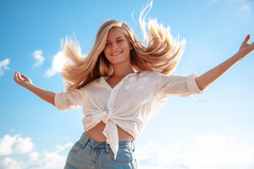 Beautiful young blonde girl with flowing hair and dimples against the blue sky and the sun, beauty and fashion