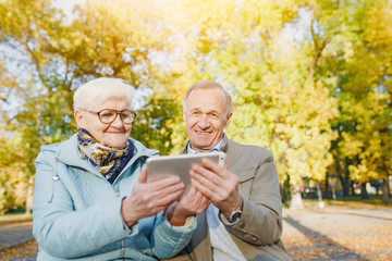 Senior couple sitting on a bench in autumn park and using digital tablet