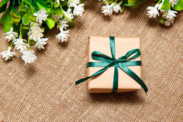 Top view of gift box in kraft paper with green satin ribbon with white flowers laying on sackcloth. Spring holidays