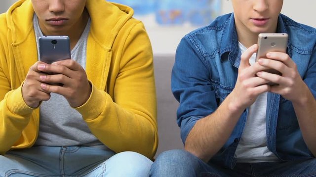 Two school friends slacking on couch playing video games on smartphones, leisure