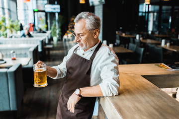 senior worker in apron holding glass of beer in pub