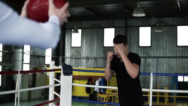 Motivated, young boxer in casual is training with coach on the ring. View from the back of the coach holding a heavy ball while guy is practicing punches and tactics steps. Old style gym on the
