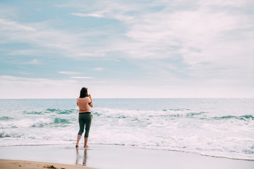 Fototapeta na wymiar Young Caucasian Woman Standing On Summer Sea Ocean Beach And Taking Photos On Smartphone.