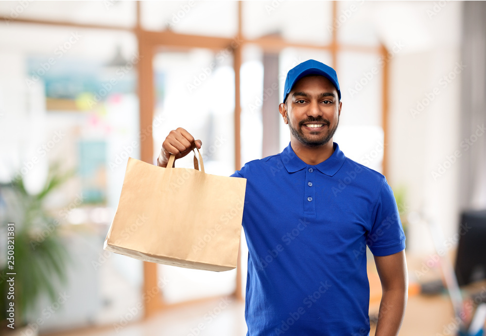 Wall mural takeaway service and people concept - happy indian delivery man food in paper bag in blue uniform over office background