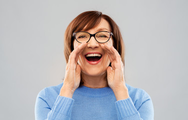announcement and old people concept - senior woman in glasses calling over grey background