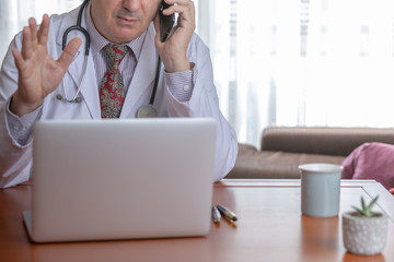 Male doctor talking over the mobile phone while looking at the laptop with an infusion beside him