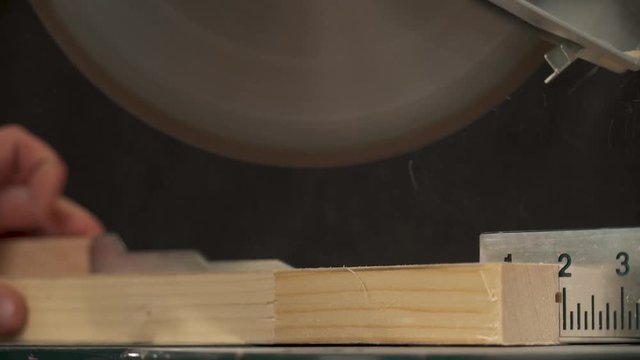 A close-up of a miter saw making a cross-cut of a work piece with sawdust flying. A hand is taking cut work piece