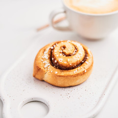 Freshly baked cinnamon roll with spices and cocoa filling and coffee or cappuccino with latte art on white serving plate over white marble background. Swedish breakfast. Close up. Selective focus.