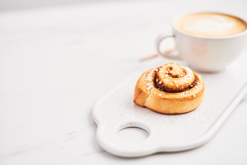 Freshly baked cinnamon roll with spices and cocoa filling and coffee or cappuccino with latte art on white serving plate over white marble background. Swedish breakfast. Copy space. Selective focus.