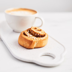 Swedish breakfast. Freshly baked cinnamon bun with spices and cocoa filling and coffee or cappuccino on white serving plate over white marble background. Close up. Selective focus.
