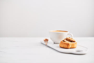 Freshly baked cinnamon roll with spices and cocoa filling and coffee or cappuccino on white serving plate on white marble table over white background. Swedish breakfast. Copy space. Selective focus.