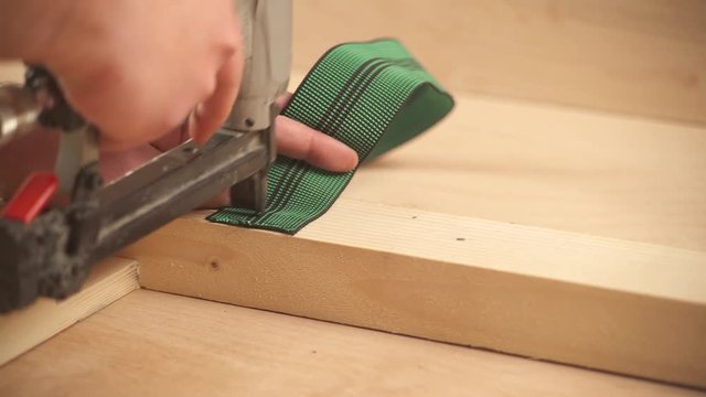 A close-up view of a staple gun attaching webbing to a wooden frame with staplers