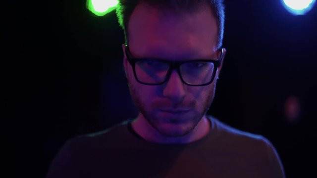 Portrait of handsome bearded guy in glasses drinking alcoholic drink from a glass standing on the background of flickering light in a nightclub.