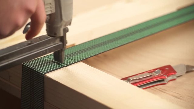 A close-up view of man's hands holding pneumatic staple gun and attaching webbing to a wooden frame. An extra webbing is cut off.
