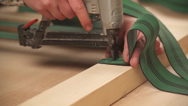 A close-up view of man's hands holding a staple gun and attaching next semi elastic band to a frame.