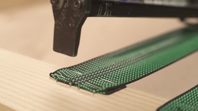 A close-up of a pneumatic staple gun attaching webbing to a wood piece with staples.