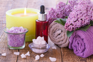 Red bottle with aromatic oil, burning candle, bowls with sea salt, lilac flowers and towels.