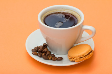 Cup of coffee with coffee beans and delicious macaron cake on peach background.