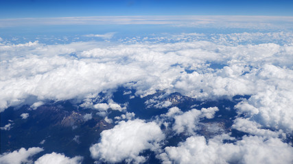 Flying over fluffy white clouds with a partial view of the rural landscape far beneath. Picturesque moving cloudscape view from airplane window.