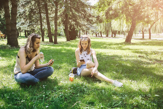 Two hipster girls are sitting on the grass with summer cocktails. Sisters are looking at their smartphones. Social networks communication. Cold non-alcoholic drinks with ice to go. Mojito and lemonade