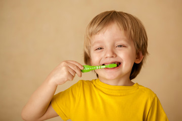 Cute little blond boy 4-5 years old in a yellow T-shirt brushes his teeth with a colorful baby toothbrush looking at the camera