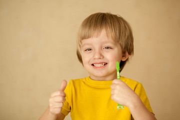 Cute little blond boy 4-5 years old in a yellow T-shirt brushes his teeth with a colorful baby toothbrush looking at the camera
