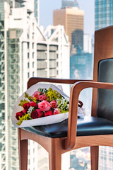 Bouquet of red and pink roses wrapped in white paper on wooden chair. Business office buildings on background behind the window. Focus on flowers. 