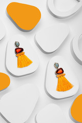 Closeup top view of two orange tassel earrings, decorated with faceted stones. The set is isolated on snowy droplike platforms. The background is decorated with snowy and flame-colored platforms. - obrazy, fototapety, plakaty