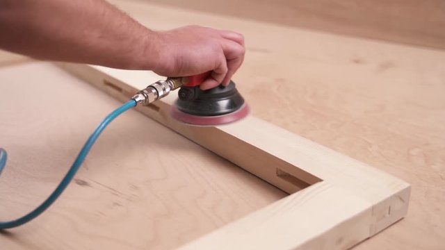 A front view of man's hand holding a pneumatic power sander and polishing a wooden frame joined by bridle joint