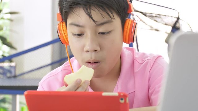 Happy asian boy playing on tablet computer with orange headphone, Teenage boy eating piece of apple and watching on tablet.