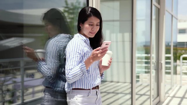 cheerful businesswoman in smart casual enjoying coffee break outdoors on sunny day. young office lady relaxing relying on window outside in office terrace. concentrated young girl holding cellphone.
