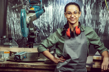 Smiling craftswoman grinding timbers with special machine. Beautiful woman wearing safety glasses. Concept of joiner's shop and woodworking. Gender equality. Male profession