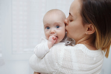 Young happy loving mother plays kisses her baby daughter in her arms against the window. Maternal care. Childcare. Close-up.