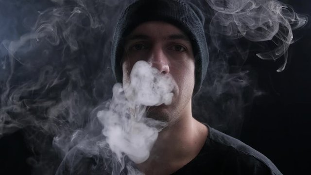 young man vaper exhaling big clouds of smoke with e-cigarette vape on black background in slow motion