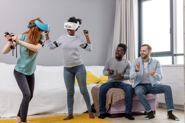 Two multiracial young women dancing wearing virtual reality glasses at home while their boyfriends...
