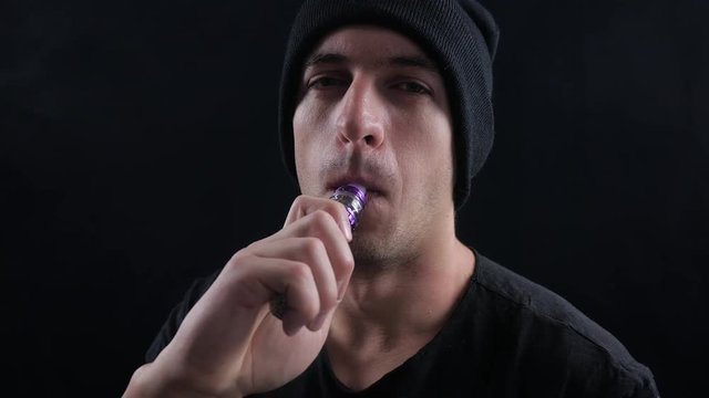 young man vaper exhaling big clouds of smoke with e-cigarette vape on black background in slow motion
