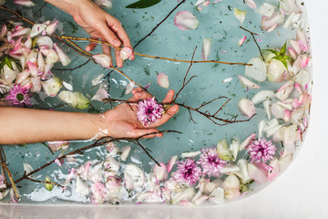 Top view of bath filled with blue bubble water, flowers, branches and petals with woman's hand, spa...