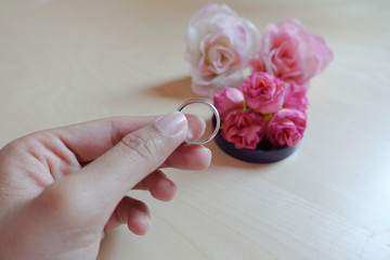 A hand holds wedding ring in front of pink fabric roses on wood table