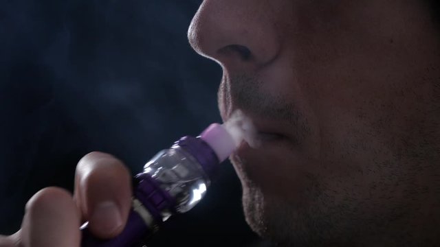 Close-up of man vaper exhaling big clouds of smoke with e-cigarette vape on black background in slow motion